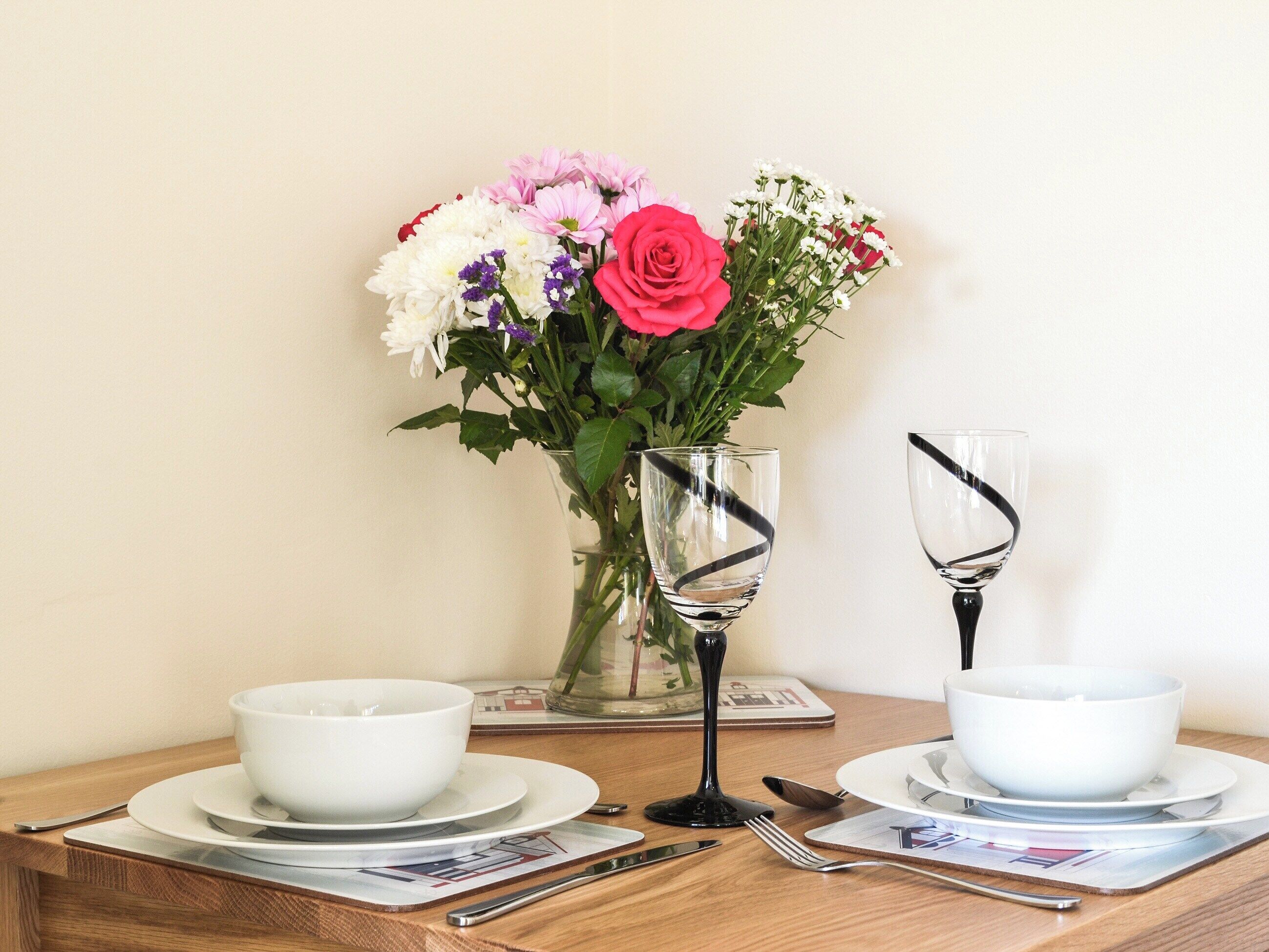A dining table set with white bowls, a glass vase with flowers, and cutlery.