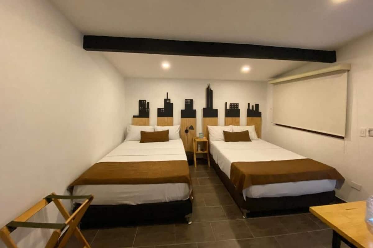 A hotel room with two beds, a wooden nightstand, a small table, and a cityscape mural on the wall.
