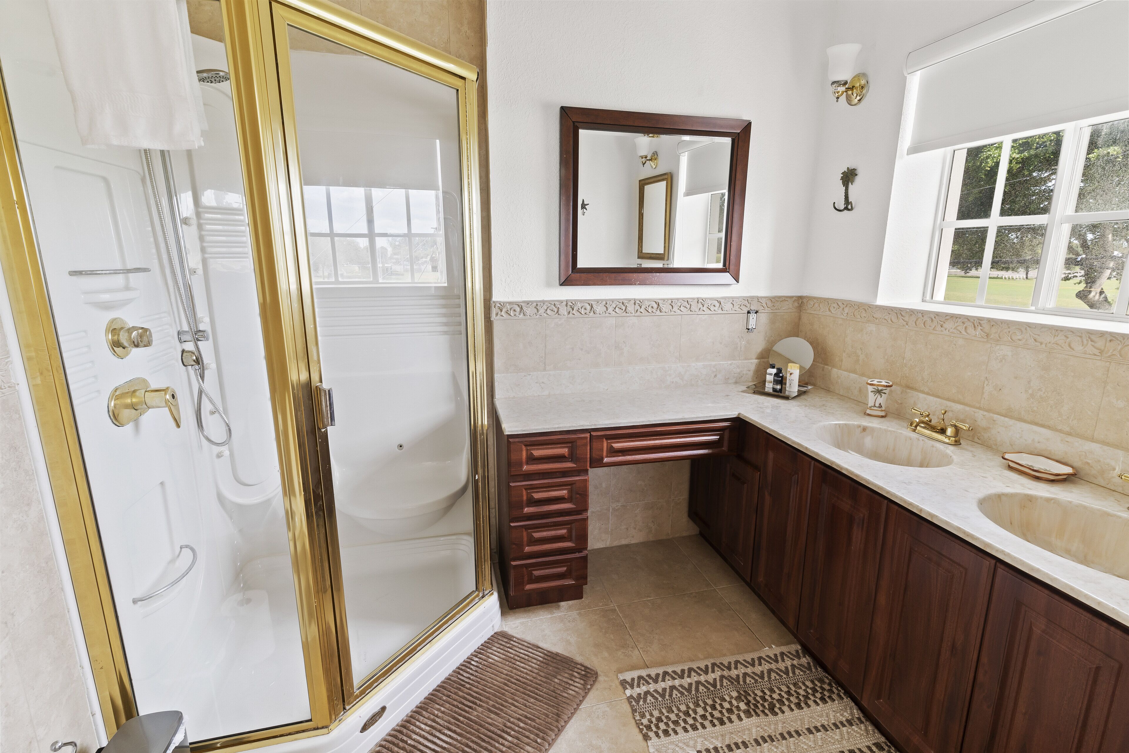 A bathroom with a walk-in shower, a large mirror, a vanity with two sinks, and a window providing a view of the outdoors.