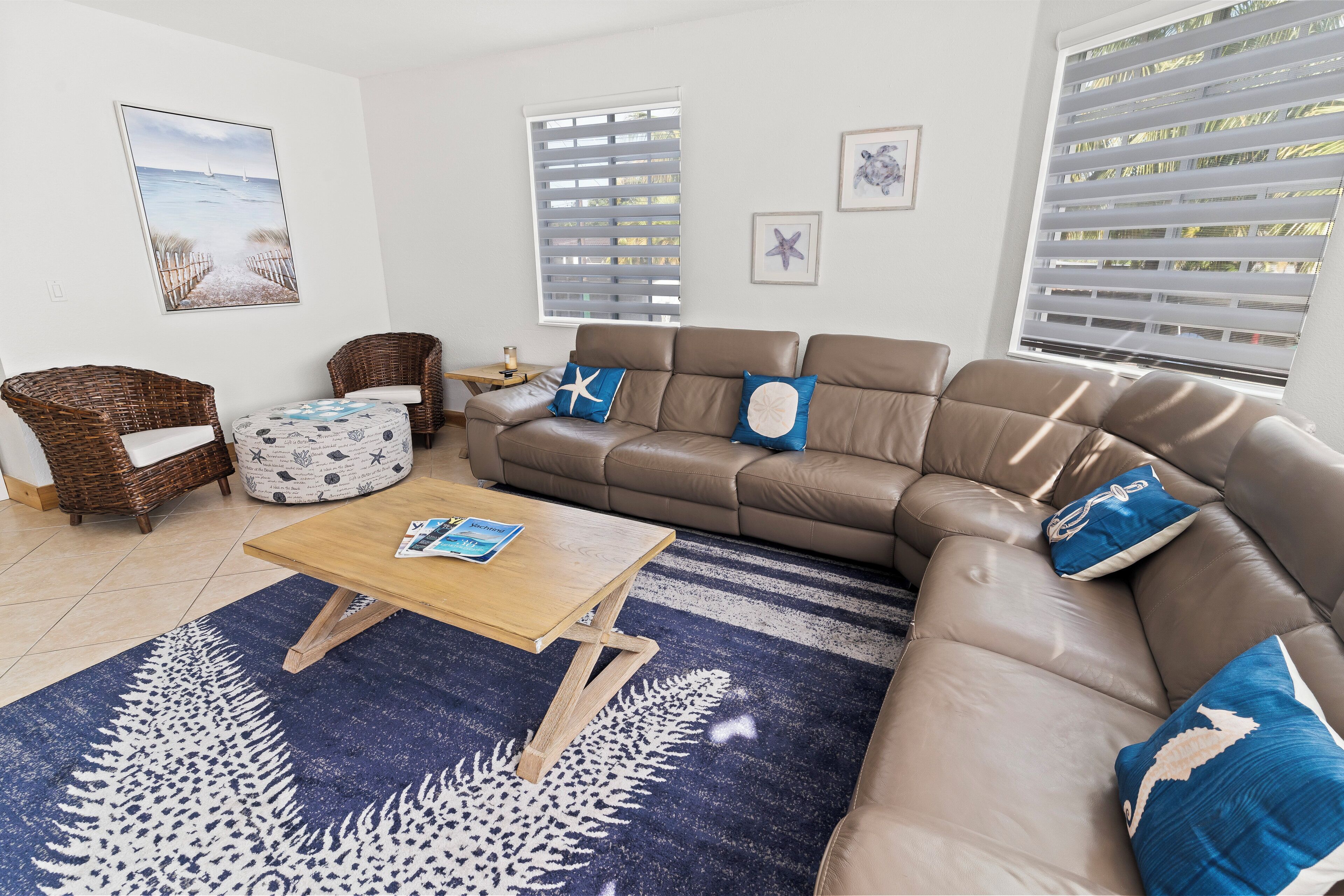 A living room with a brown leather sofa, two wicker chairs, a wooden coffee table, and a blue patterned rug.