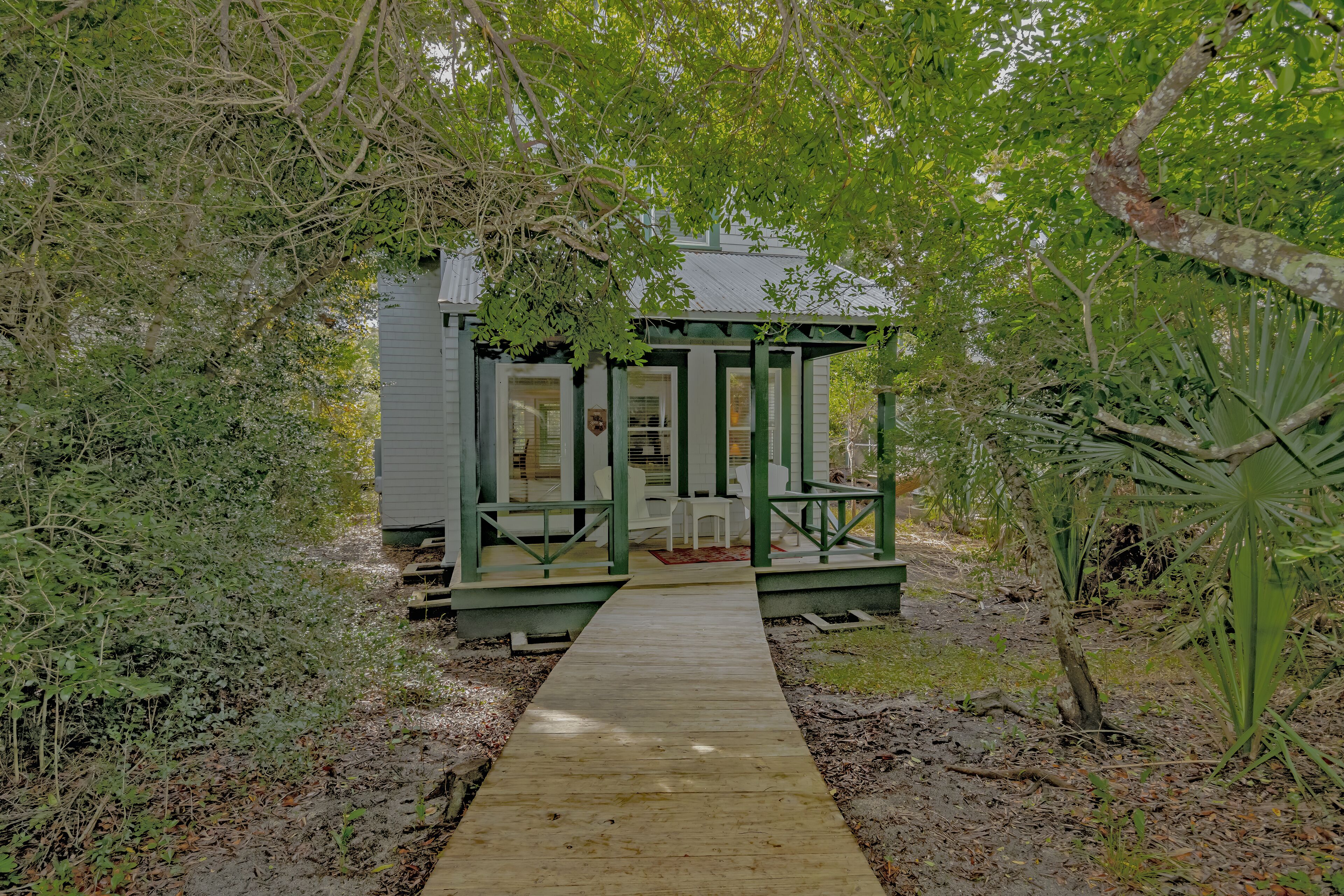 A wooden pathway leading to a small structure with a green roof and white walls, surrounded by dense foliage.