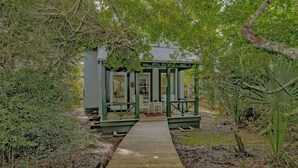 A wooden pathway leading to a small structure with a green roof and white walls, surrounded by dense foliage. - Tranquil Family Home located in Surfman's Walk; private crofter with shoals access (Bald Head Island)
