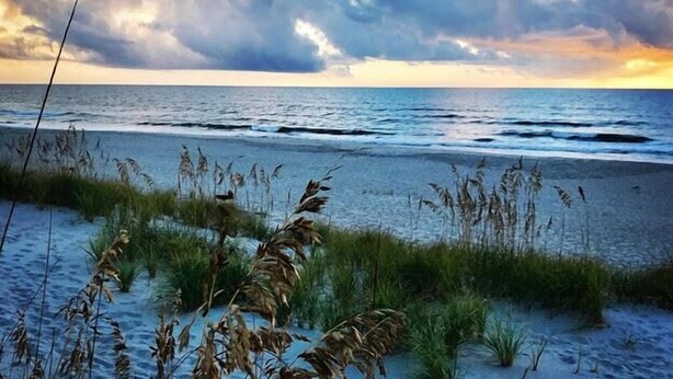 A beach with waves, grassy dunes, and a cloudy sky. - Tranquil Family Home located in Surfman's Walk; private crofter with shoals access (Bald Head Island)