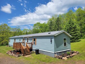 A single-story house with a front porch and a small deck, surrounded by a stone border and a grassy yard. - Beautiful 4 season getaway in the scenic northwoods of the Western UP! (Bessemer)