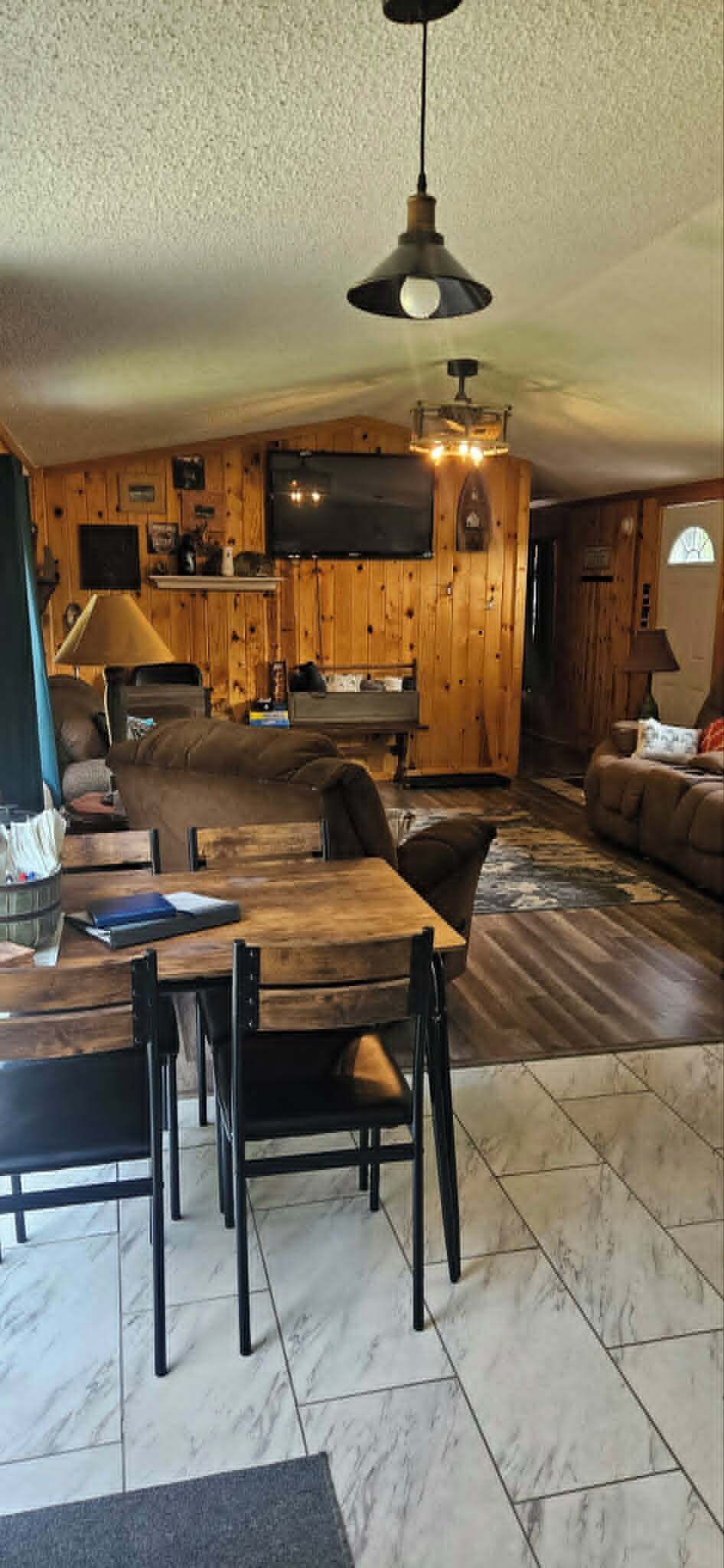 A cozy living room with a wooden wall, a dining area with a table and chairs, and a television mounted on the wall.