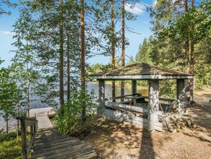 Ein hölzerner Pavillon mit Bank und Tisch, umgeben von hohen Bäumen und einem Gewässer.