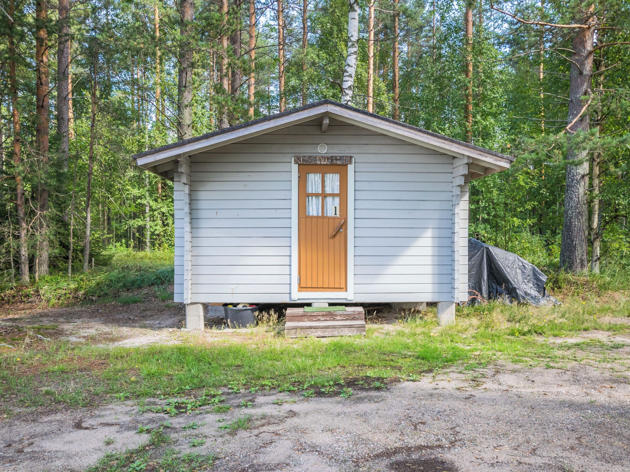 A small, single-story cabin with a wooden door and a small porch, surrounded by a forested area.