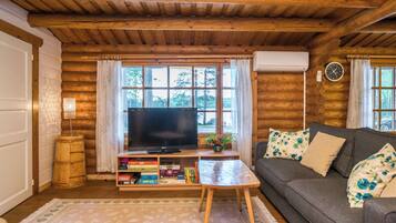 Interior acogedor de una cabaña de madera con techo de madera, un televisor sobre un soporte de madera, un sofá con cojines estampados y una mesita con una lámpara.