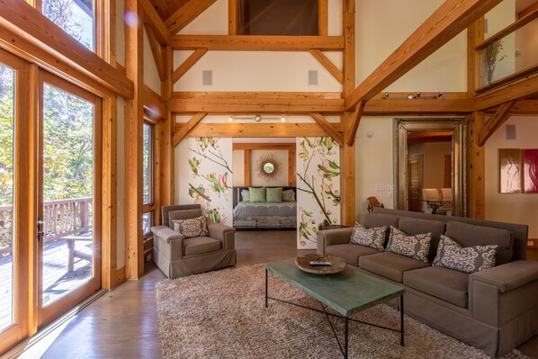 A spacious living room with a large sofa, a coffee table, and a fireplace. There is a balcony with wooden railings and a view of trees.