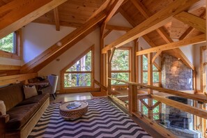 A cozy loft-style room with a stone fireplace, wooden beams, and large windows offering a view of the forest.