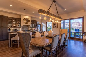 A spacious dining area with a large wooden table and chairs, a hanging light fixture, and a view of the outdoors through large windows.