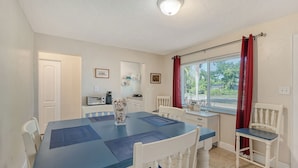 Un coin repas avec une table bleue et des chaises blanches, un petit espace cuisine équipé d’un four à micro-ondes, et une fenêtre agrémentée de rideaux rouges.