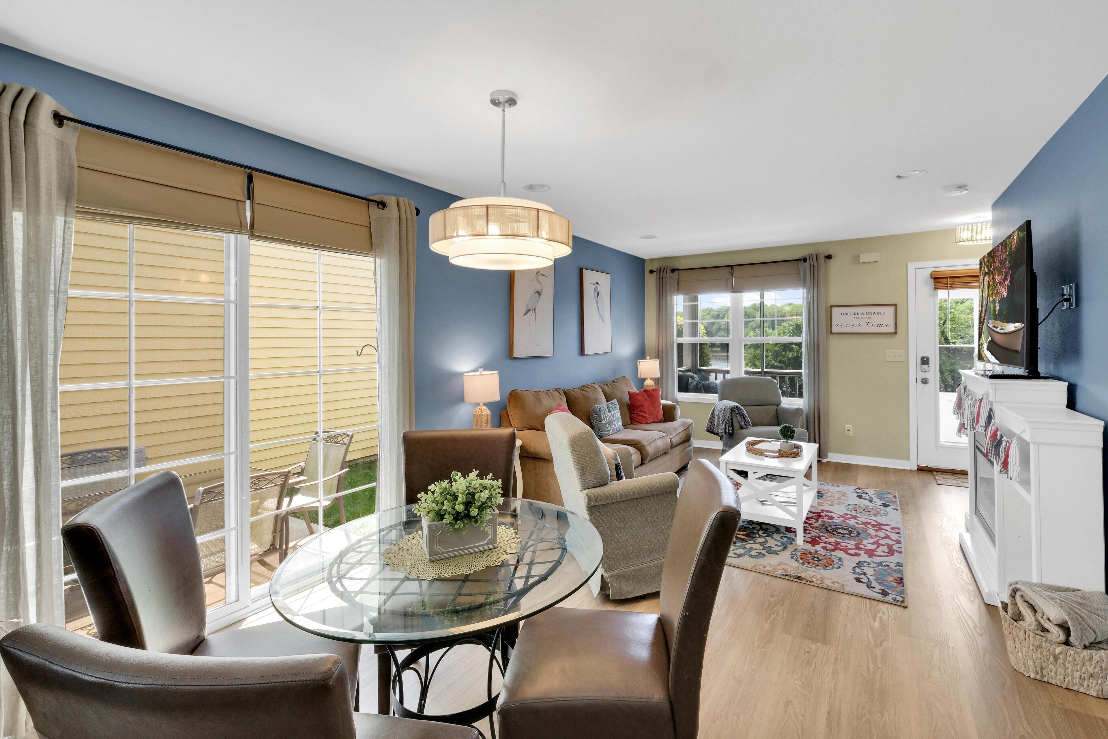 A spacious living room with a glass dining table, comfortable seating, and a large window with blinds.