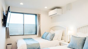 Ein Hotelzimmer mit zwei Betten, einem an der Wand befestigten Fernseher, einem Fenster mit Vorhängen und einer Klimaanlage.