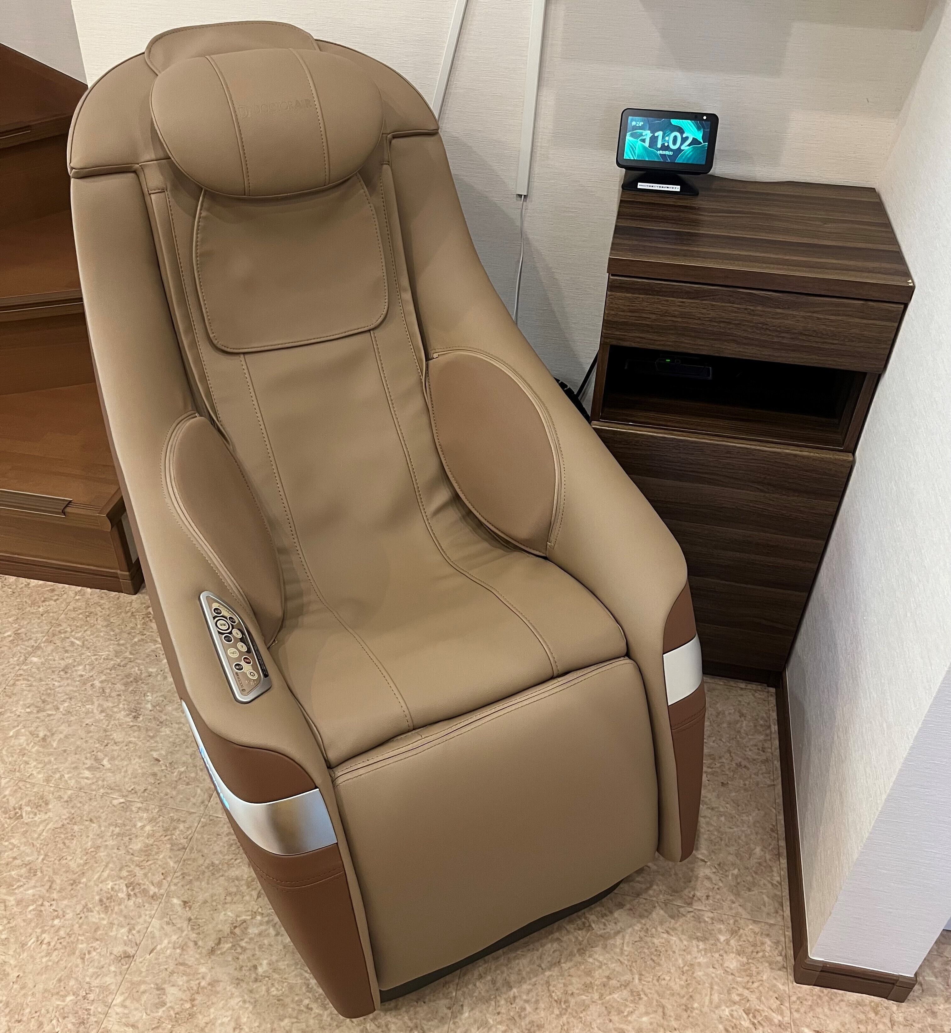 A massage chair with a control panel, positioned next to a wooden nightstand with a digital clock.