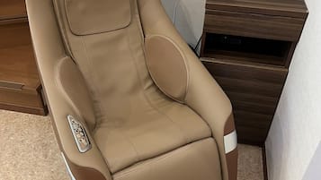 A massage chair with a control panel, positioned next to a wooden nightstand with a digital clock.