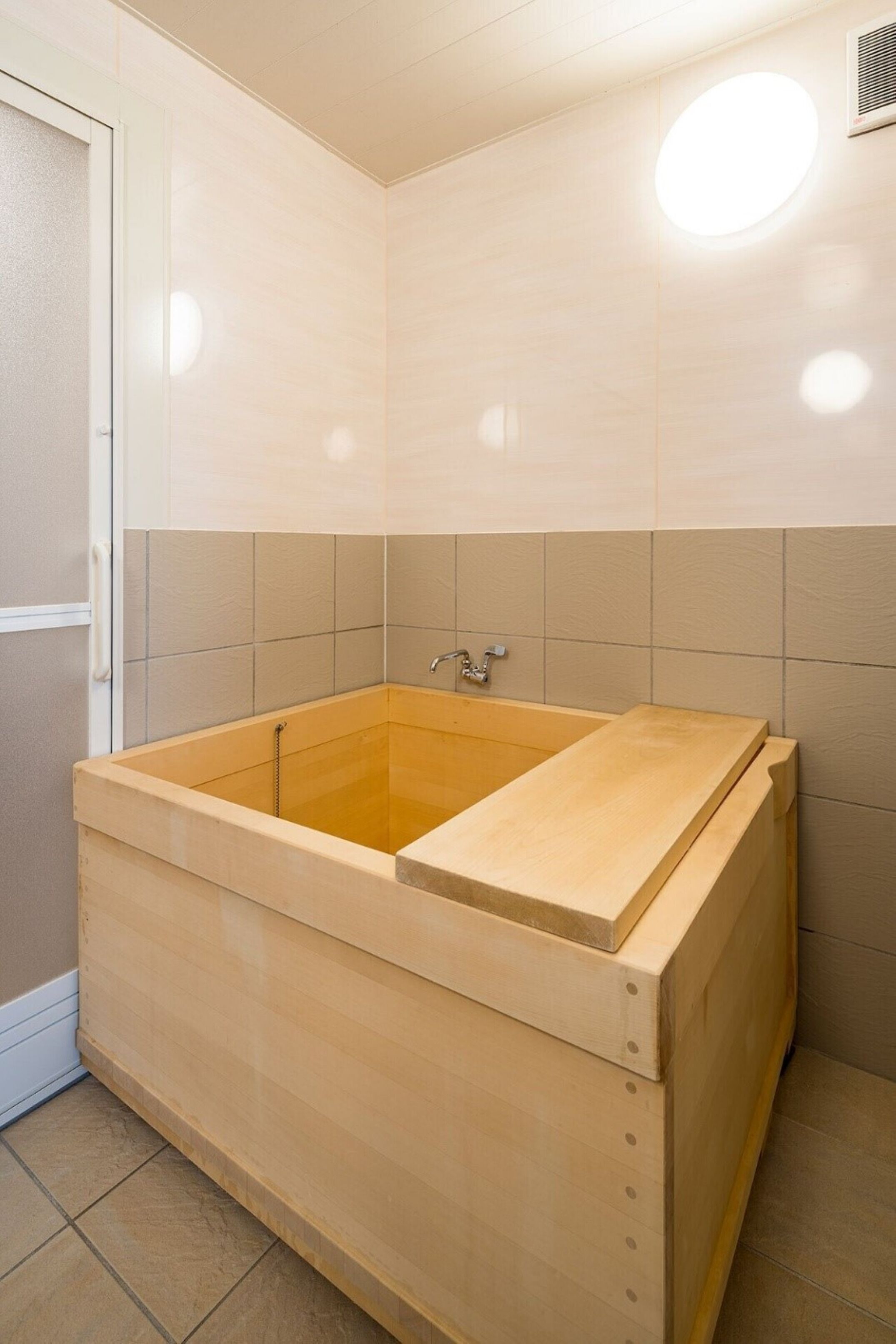 A wooden bathtub with a built-in seat, a showerhead, and a wall-mounted light fixture.