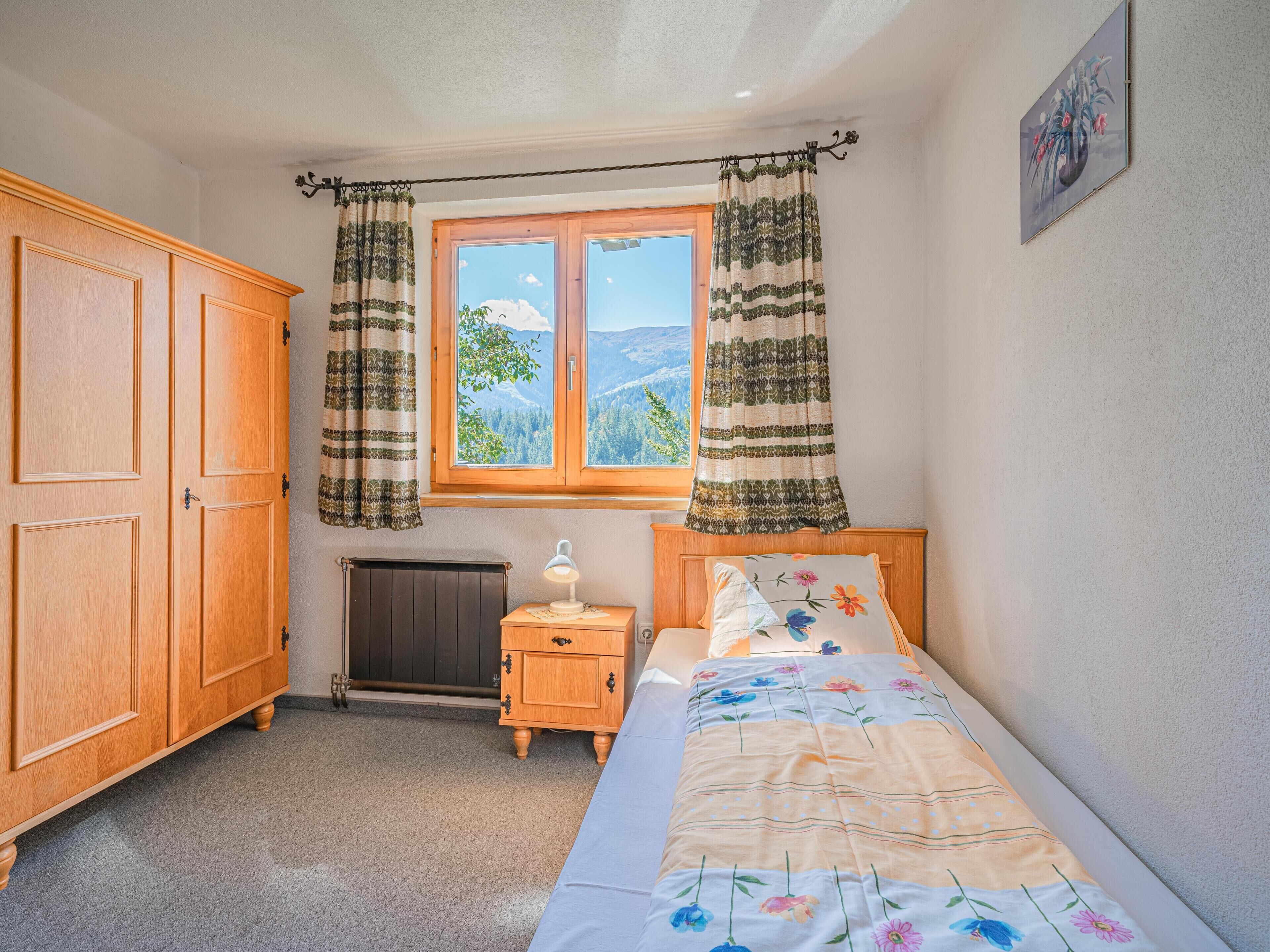 A bedroom with a wooden wardrobe, a bedside table with a lamp, a bed with floral bedding, and a window with curtains.