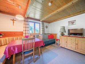 A cozy room with a wooden ceiling, a cross on the wall, a window with a view of trees, a sofa, a TV, and a table with chairs.