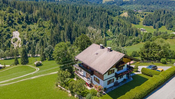 A two-story house with a sloped roof, surrounded by lush green fields and dense forest. - Sunny holiday home with open views (Westendorf)