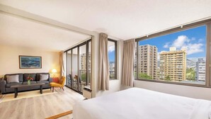 Une chambre d’hôtel avec un lit, un canapé, une petite table, une chaise, un tableau et une grande fenêtre donnant sur la ville.