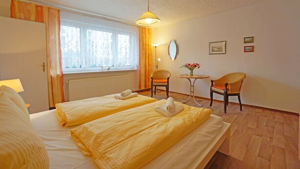 Une chambre d’hôtel avec deux lits, une petite table et une chaise, une fenêtre avec des rideaux et un mur orné de tableaux.
