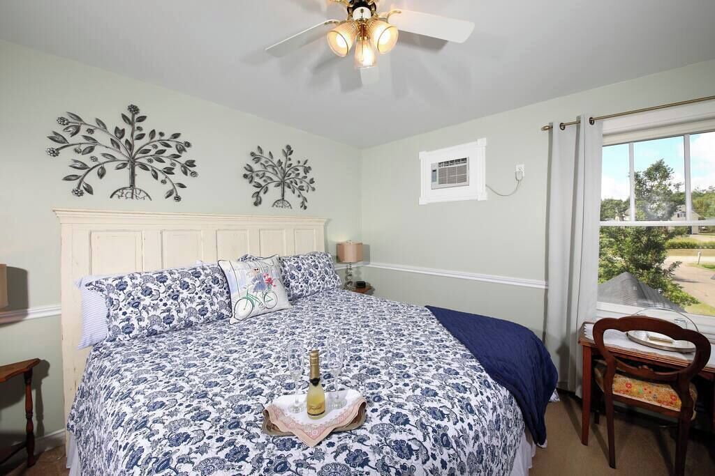Une chambre à coucher comprenant un lit, un ventilateur de plafond, des œuvres d’art décoratives au mur, une fenêtre avec des rideaux et une vue sur l’extérieur.
