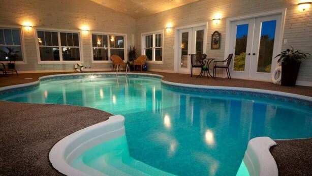 En indendørs swimmingpool med klart, blåt vand, omgivet af en træterrasse og en lille patio med stole og et bord.
