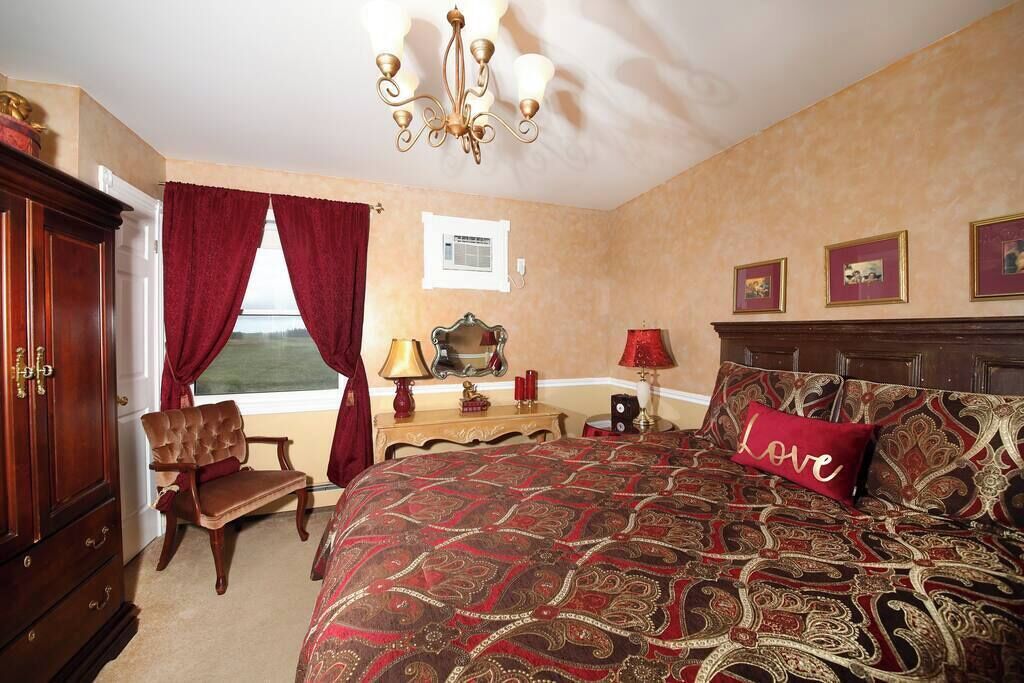 Quarto com cama grande, guarda-roupa de madeira, cadeira, escrivaninha com luminária e janela com cortinas.