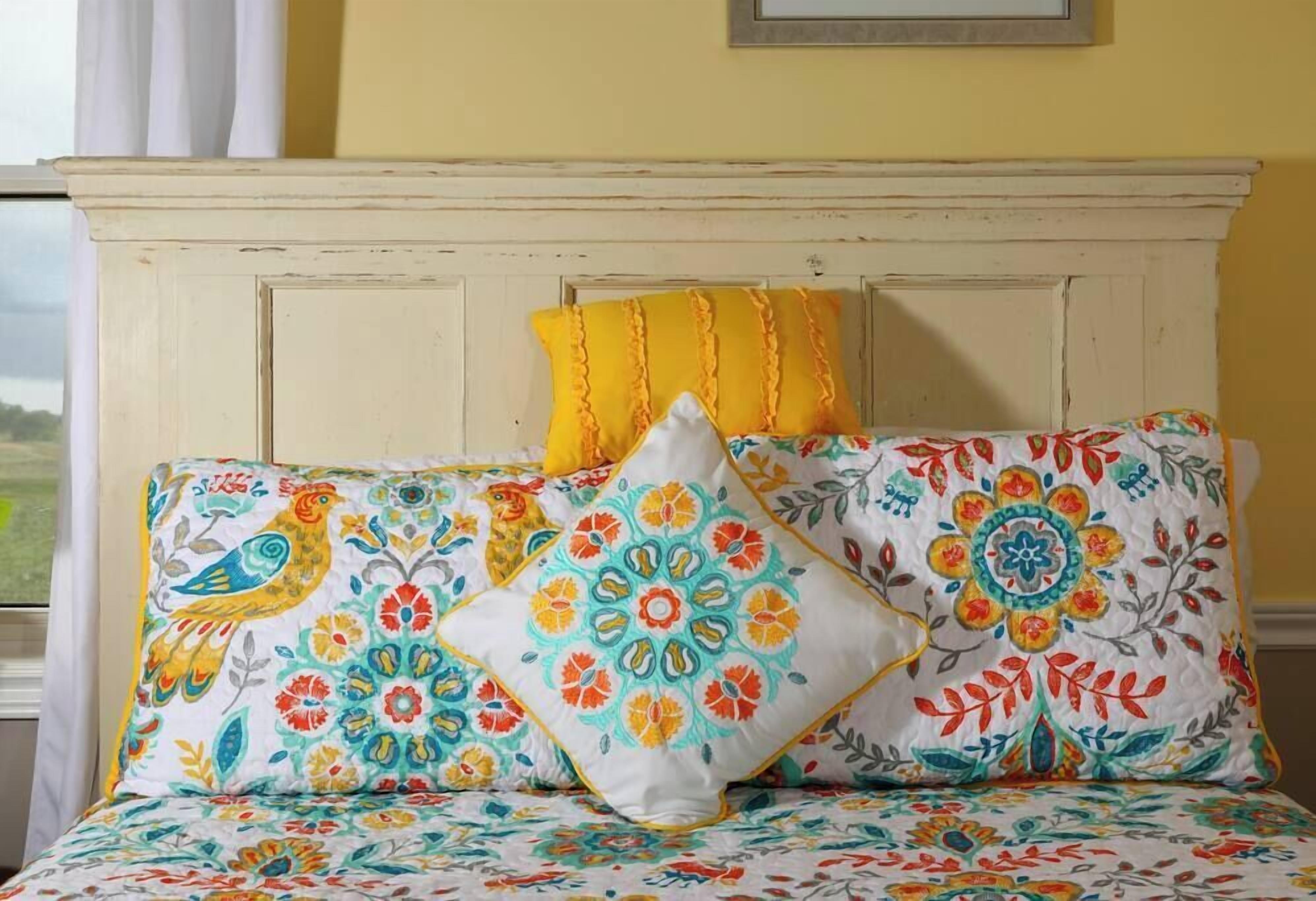 Un lit avec une literie aux motifs floraux colorés et des coussins décoratifs, une tête de lit blanche et une fenêtre avec des rideaux.