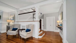 A spacious living room with a wooden floor, two armchairs with blue cushions, a white fireplace, and a staircase leading to an upper level. - Magnificent Ferncrest - Towering Home on the Coast (Groton)
