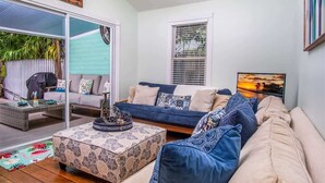 A living room with a sofa, coffee table, and a TV. There is a window with blinds and a door leading outside. - Runaway Bills (Key West)