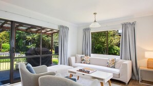 Ein modernes Wohnzimmer mit einem weißen Sofa, zwei Sesseln, einem Couchtisch und einem Schachbrett. Große Fenster mit grauen Vorhängen bieten Blick auf einen Garten und eine Grillplatz.