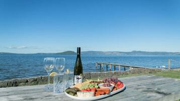 A wooden table with a cheese platter, wine glasses, and a bottle of wine by a waterfront.
