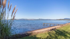 A lakeside view with a wooden dock extending into the water, surrounded by grassy areas and tall grasses. - Front Row on the Lake | Jetty, Kayaks & Room to Spread Out (Rotorua)