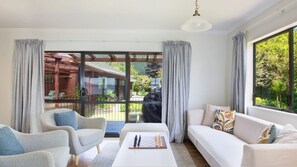 A modern living room with a sofa, armchairs, and a coffee table. There is a large window with curtains, and a view of the outside. - Front Row on the Lake | Jetty, Kayaks & Room to Spread Out (Rotorua)