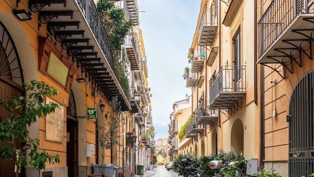A narrow street with buildings on both sides, featuring balconies with plants and outdoor seating.