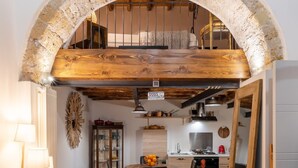 A cozy interior with a wooden dining table, a yellow armchair, and a kitchen area visible through an arched doorway.
