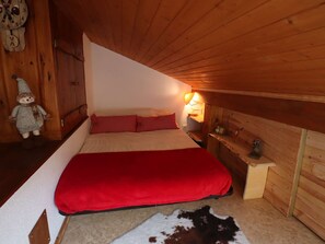 Une chambre confortable avec un plafond en pente, un lit avec une literie rouge, une petite table de chevet avec une lampe et un mur en bois orné d’une horloge.