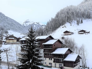 Uma aldeia montanhosa coberta de neve, com edifícios de vários andares, incluindo um hotel com varandas e uma estância de esqui ao fundo.