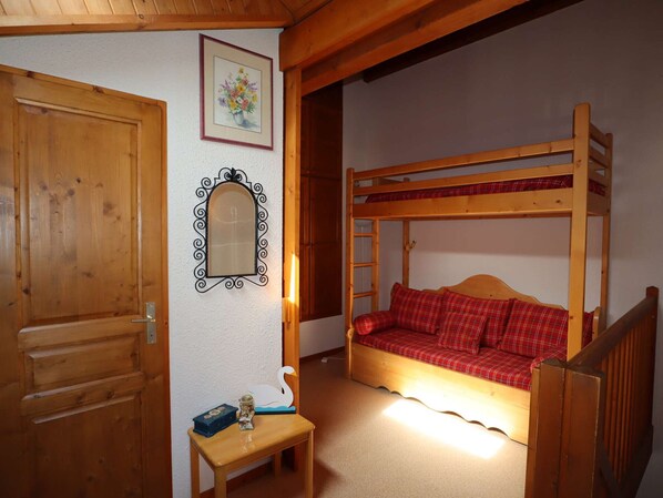 A wooden bunk bed with red plaid bedding, a small table with a swan decoration, a framed mirror, and a door. - Stunning 6-Person Duplex - Ski, Summer Fun, WiFi, Garage, Pool (Arêches-Beaufort)
