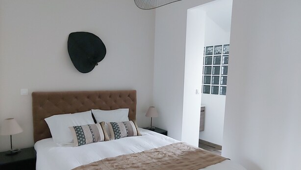 A bedroom with a large bed, a headboard, a hanging light fixture, and a window with blinds.