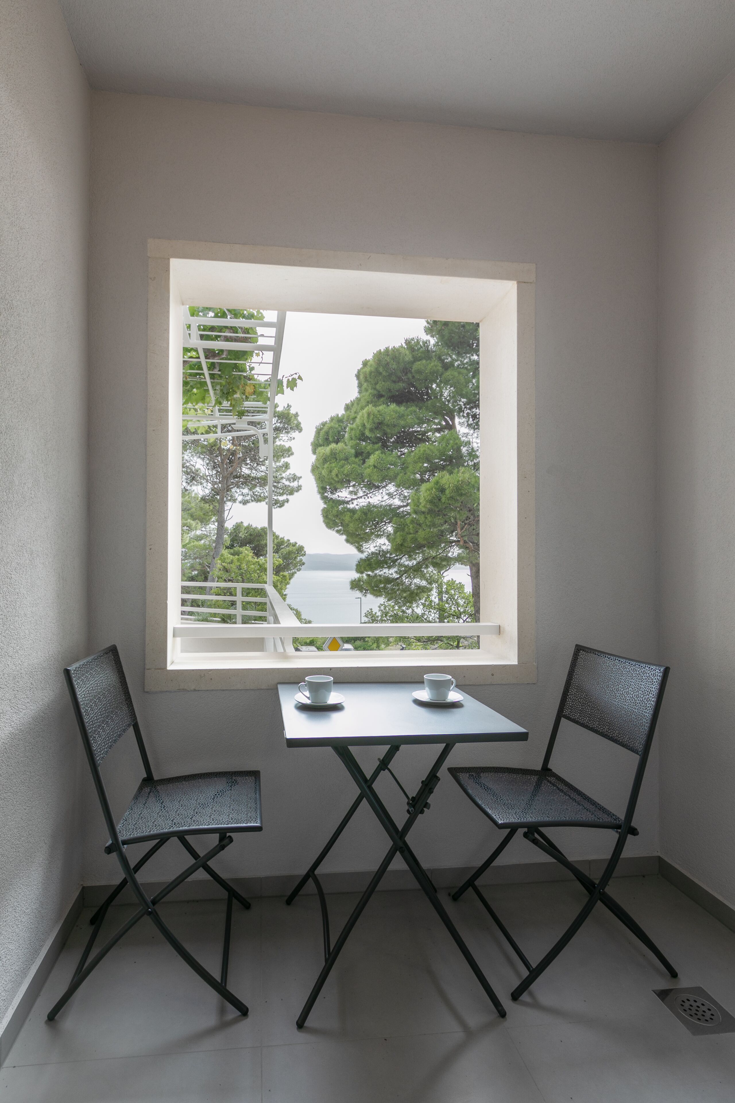 A small room with a window overlooking a body of water and trees. There is a table with two chairs and two cups on it.