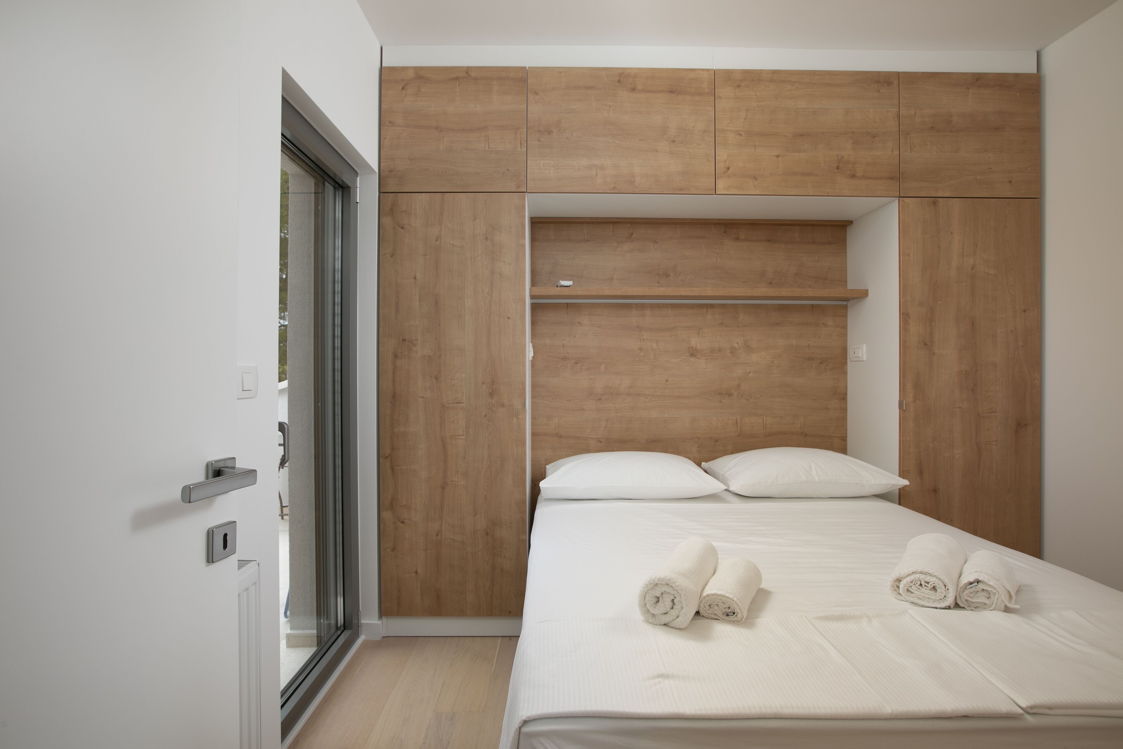 Une chambre moderne avec une tête de lit en bois, un lit recouvert de linge de maison blanc et une porte en verre donnant sur un balcon.