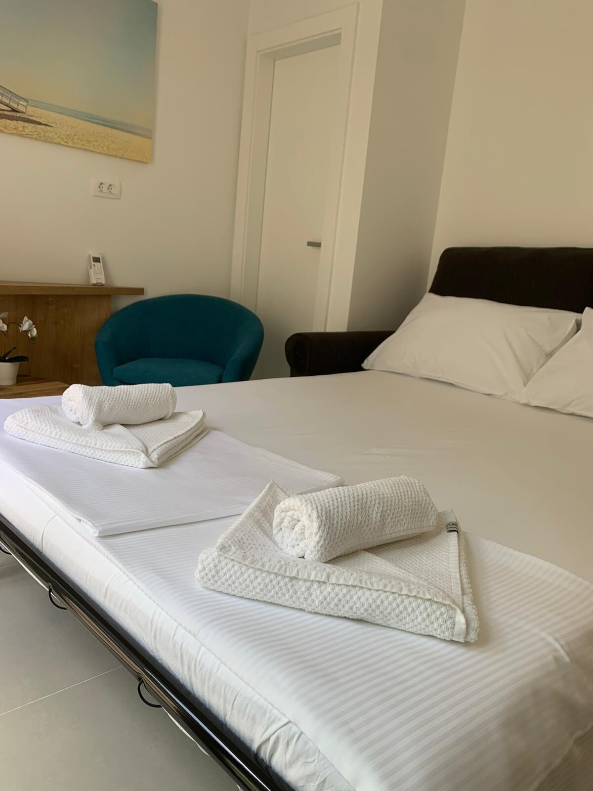 A hotel room with a bed, a teal chair, a wooden dresser, and a painting on the wall.