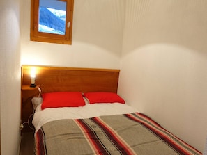Una habitación pequeña con cama, mesita de noche, lámpara y ventana con vistas a un paisaje nevado.