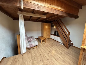 A loft bedroom with a wooden ceiling, a bed with patterned bedding, a wooden bench, a wardrobe, and a staircase.