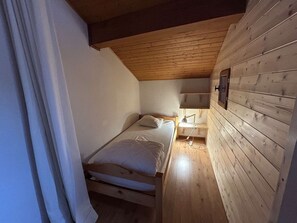 A small, cozy room with a wooden ceiling, a single bed, a bedside table with a lamp, and a wall-mounted shelf.