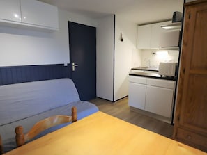Habitació petita amb un llit, una taula de fusta, una cuineta amb fogó i una porta.