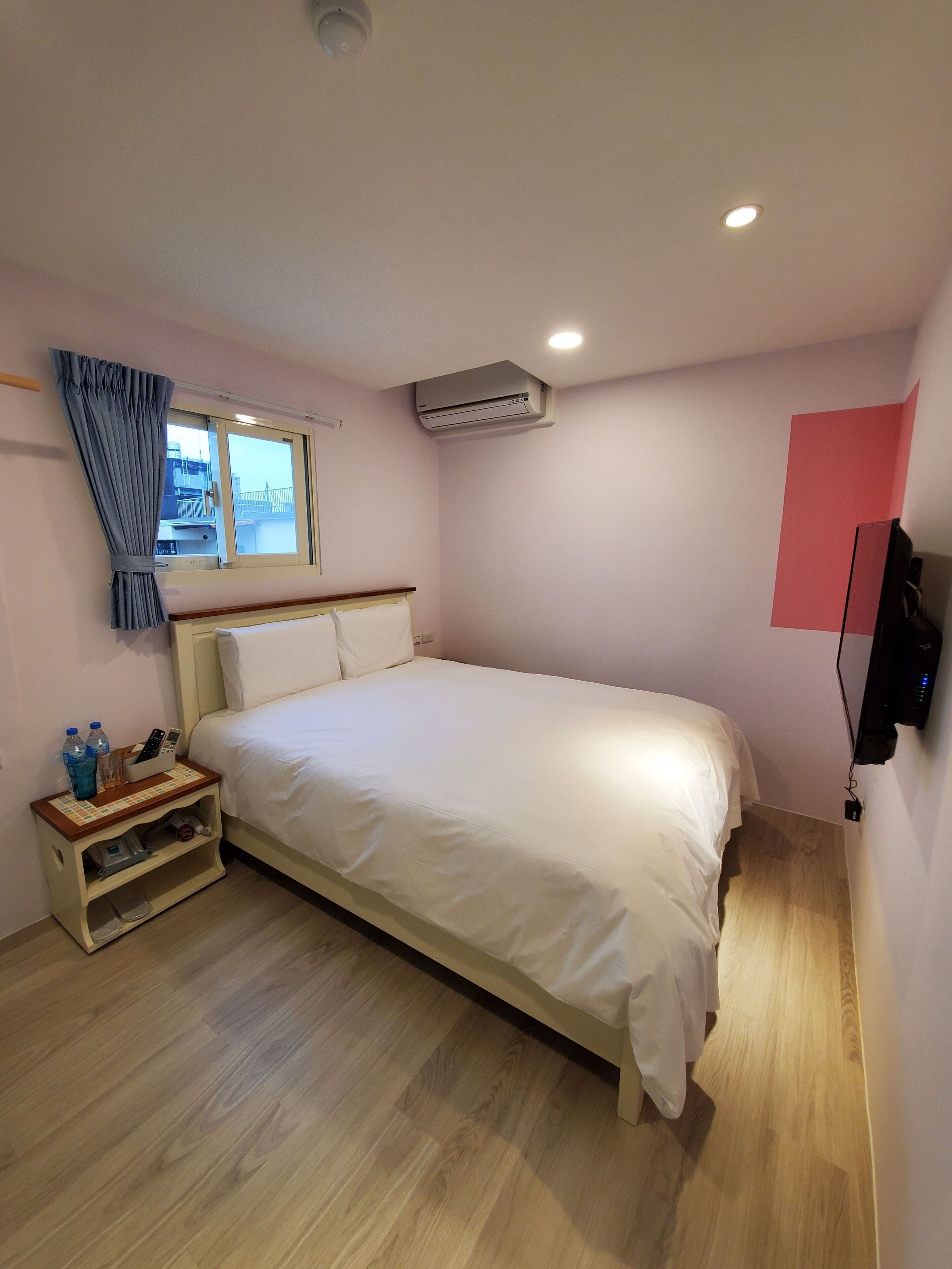 A bedroom with a bed, a bedside table with a remote control, a water bottle, and a small shelf.